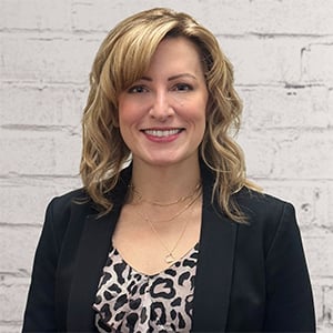 Woman with shoulder-length wavy blonde hair wearing a black blazer over a leopard print top against a white brick wall.
